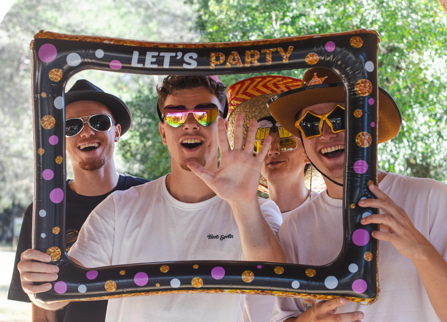 Quatre amis posent derrière un cadre gonflable « Let's party », avec des lunettes fantaisie et des chapeaux.