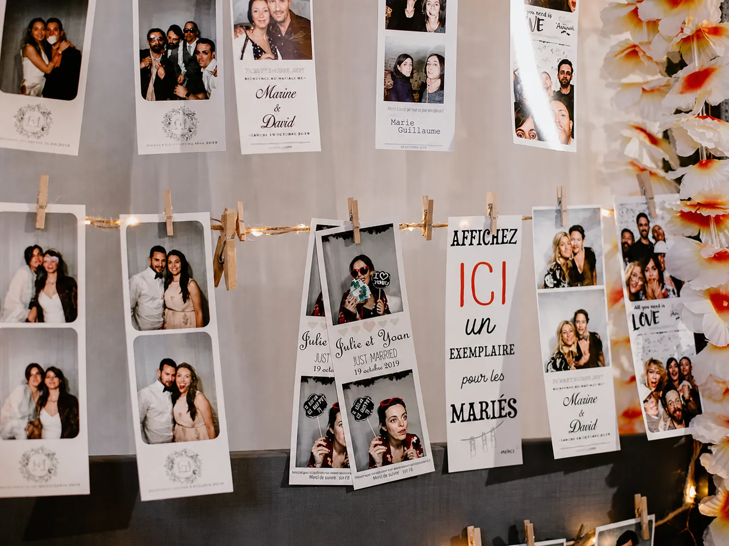 Des bandes photo suspendues à un fil avec des pinces à linge et une guirlande lumineuse, à côté d'une pancarte « Affichez ici un exemplaire pour les mariés ».